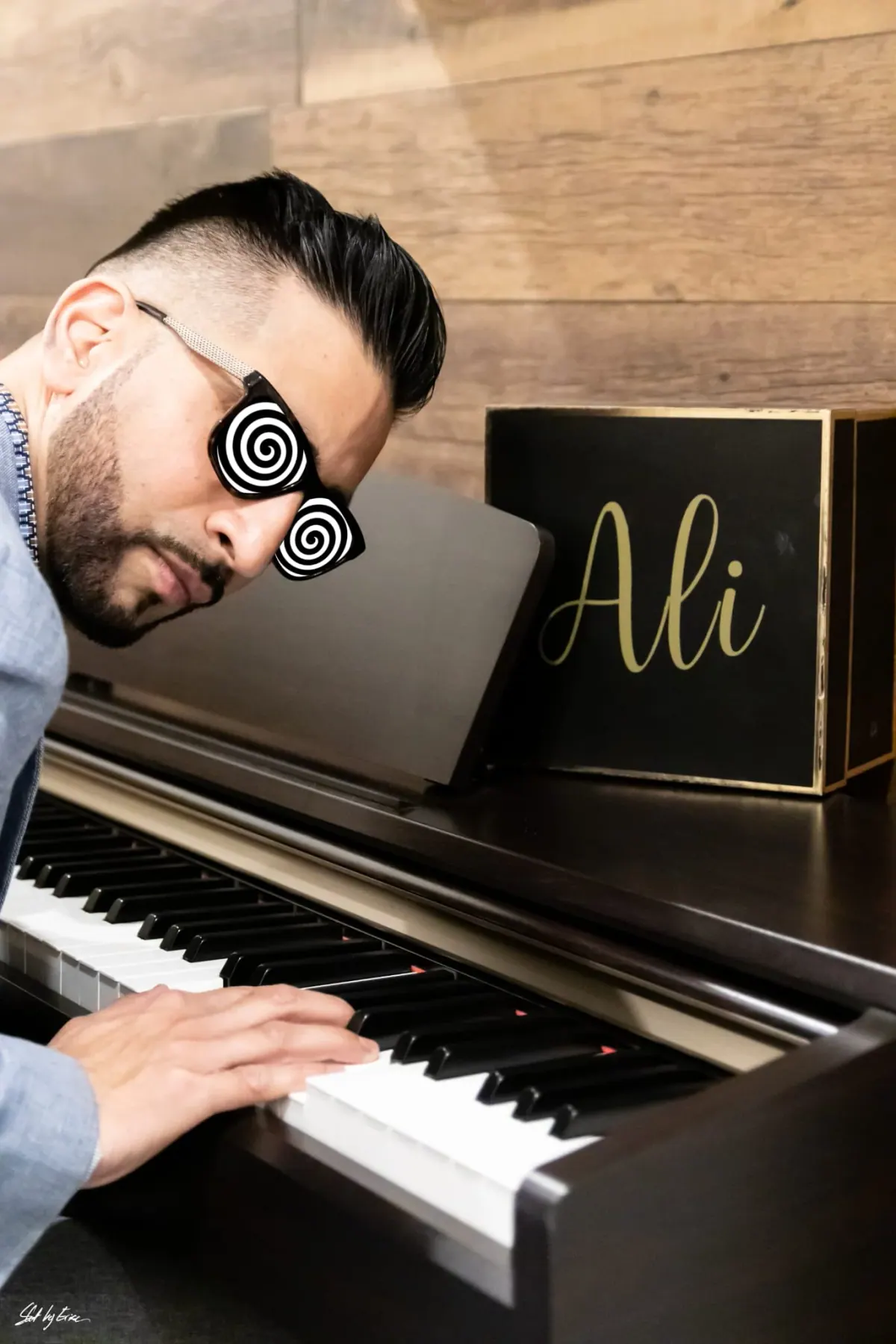 Ali in spiral novelty sunglasses leaning over a piano keyboard, beside a gold Ali sign