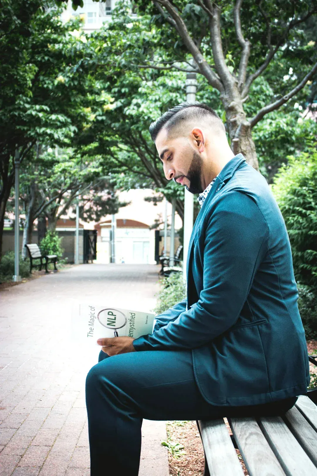 Ali on a park bench in a teal suit, reading a book about NLP
