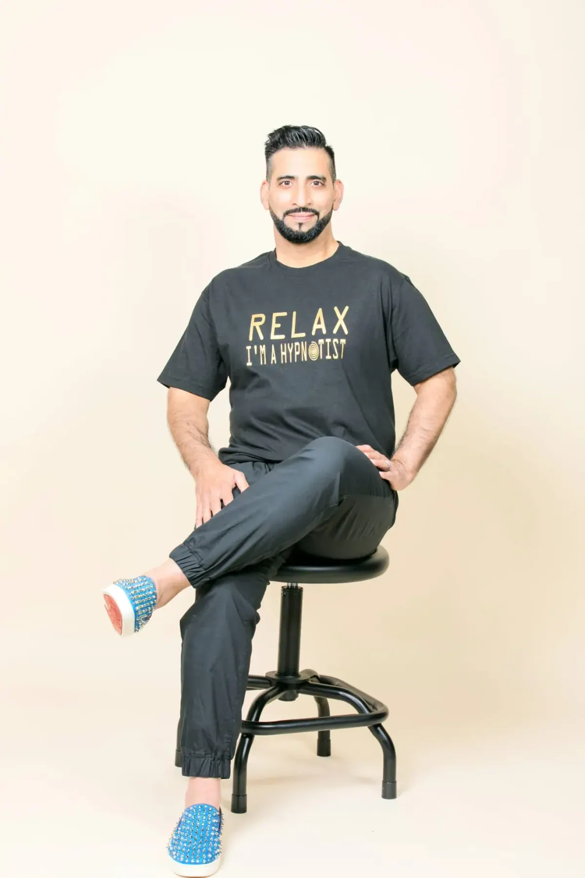 Ali seated on a stool against a plain backdrop in a tee reading Relax, I'm a hypnotist